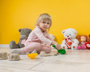 A two-year-old girl in pink suit is sitting on the floor in the playroom and playing with beige kinetic sand