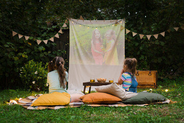 Open air cinema. Backyard Family outdoor movie night with kids. Sisters spending time together and watching cimema at backyard. DIY Screen with film. Summer outdoor weekend activities with children.