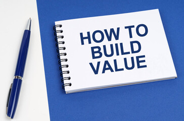 On a blue-white surface lies a pen and a notebook with the inscription - How To Build Value