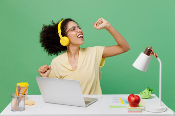 Young employee business woman of African American ethnicity in shirt sit work white office desk with pc laptop wear headphones listen music sing song in microphone isolated on plain green background