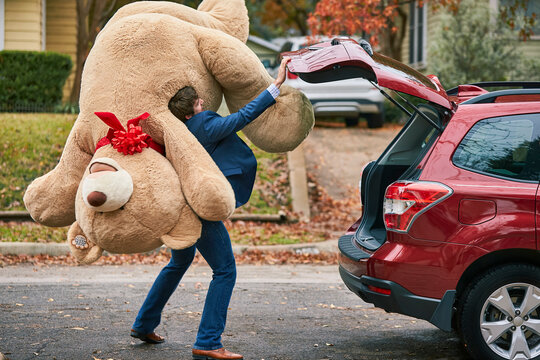 Wide shot of a man holding oversized teddy bear over his shoulder, closing the trunk of his car.