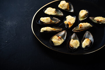baked mussels in shells with creamy sauce poured with oil on a black plate on a dark background