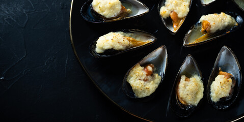 baked mussels in shells with creamy sauce poured with oil on a black plate on a dark background