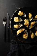 baked mussels in shells with creamy sauce poured with oil on a black plate on a dark background