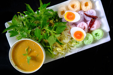 Top view Colorful Fermented Rice Flour Noodles with Fish Curry Sauce in a bowl, vegetables and boiled egg with spicy chili served on white dish and black background, popular food in Thailand
