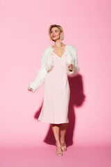 full length of stylish young woman in slip dress and faux fur jacket smiling on pink.