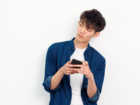 Portrait of handsome Chinese young man with curly black hair in blue shirt posing against white wall background. Looking away with mobile phone in both hands, front view studio shot.