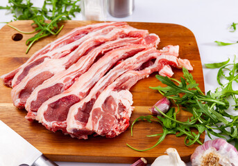 Raw lamb chops on wooden cutting board with garlic and herbs, over white background.