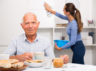 Portrait of elderly man at tea table, young woman cleaning furniture at home kitchen