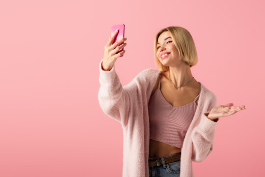 happy young woman in soft cardigan taking selfie isolated on pink.