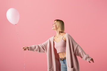 cheerful young woman in cardigan looking at balloon isolated on pink.