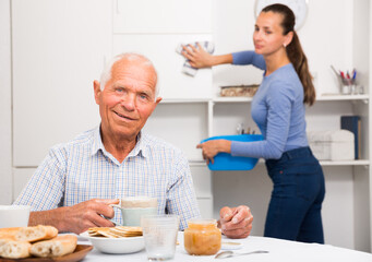 Fototapeta na wymiar Elderly man drinkimg tea, daughter washing furniture kitchen at home interior