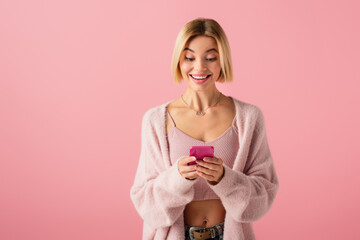 happy young woman in soft cardigan using smartphone isolated on pink.