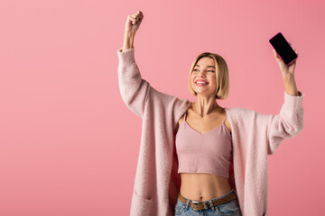 happy young woman in soft cardigan holding smartphone with blank screen isolated on pink.