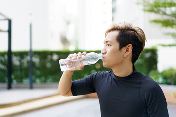 Active Asian sportsman relaxing and drinking a water in bottle after outdoor running or workout.≈