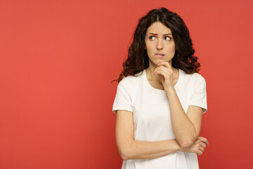Serious doubtful italian woman looking up thinking pensive and confused. Young curly girl in white outfit isolated on red background. Concerned upset female in 30s bite lips in doubts and thoughts - obrazy, fototapety, plakaty