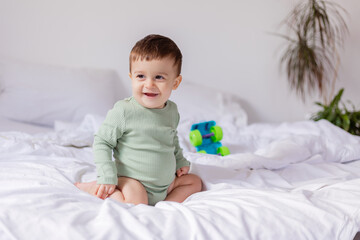 baby in a green cotton bodysuit is sitting on a white bed with a colored toy car. child is playing