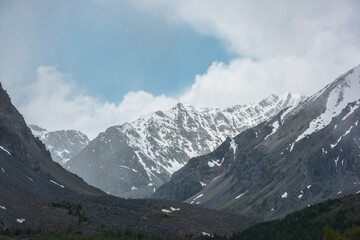 Awesome landscape with high snowy mountain range with sharp rocks in cloudy sky. Dramatic view to snow mountains in changeable weather. Atmospheric mountain scenery with white snow on black rocks.