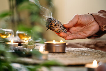 Woman hands burning white sage, before ritual on the table with candles and green plants. Smoke of...
