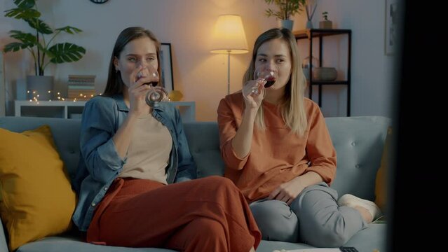 Cute young women watching TV pointing at screen and talking drinking wine in the evening in dark room. Entertainment and friends concept.