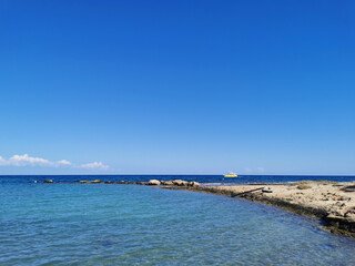 A small beach in the bay of the Mediterranean Sea with clear water and a rocky bottom against the background of stones, a yellow boat and a blue sky.