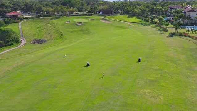 Aerial drone view of two golf cars and two people who walk in a golf course with a luxury resort in background, Dominican Republic