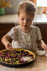 a little blonde cheerful girl cooking and decorating a pie fruits and berries on a wooden table in the kitchen with natural light coming through a window