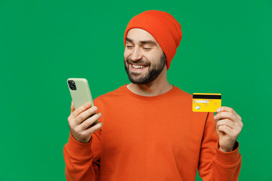 Young Smiling Happy Cool Man 20s Wear Orange Sweatshirt Hat Using Mobile Cell Phone Hold Credit Bank Card Doing Online Shopping Order Delivery Booking Tour Isolated On Plain Green Background Studio