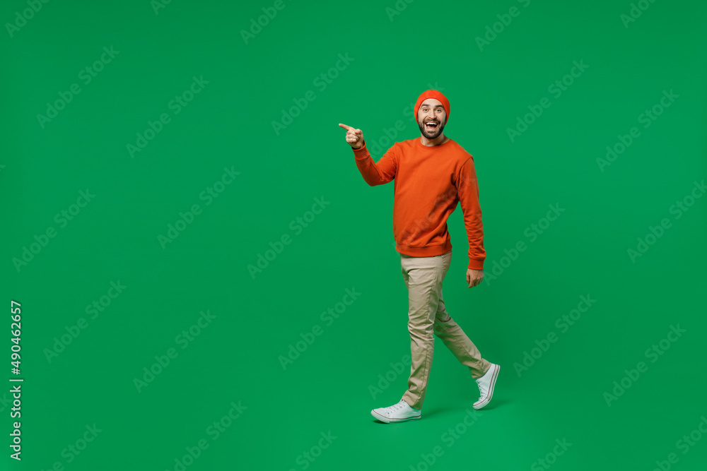 Sticker Full body young smiling happy cheerful surprised man 20s wear orange sweatshirt hat goign walking strolling point index finger aside on workspace isolated on plain green background studio portrait