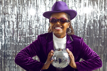 Positive young dark skinned woman in velvet suit holding disco ball with both hands. Happy young woman in hat, purple jacket and party glasses stands on background of foil silver curtain.