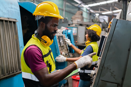 Engineering male african american workers wear soundproof headphones and yellow helmet holding tablet working at operating CNC machine. work factory industrial concept.