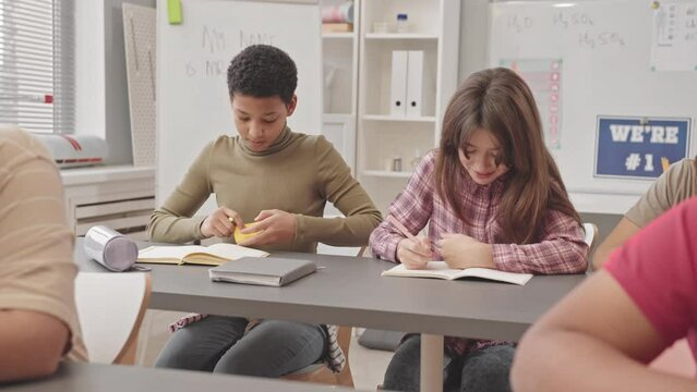 Medium slowmo of African American school girl trying to cheat during Science exam while teacher walking along desks in modern classroom