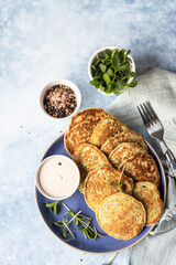 Vegetable fritters or pancakes made of zucchini, broccoli or spinach on plate with microgreen and dip, blue background. Healthy vegetarian food. Top view.