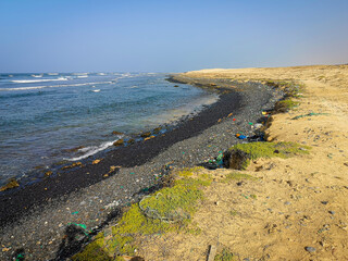 Black pebbles and garbage on the seaside, Cape Verde, Africa. Bright landscape, wavy ocean, clear blue sky. Selective focus on the details, blurred background.