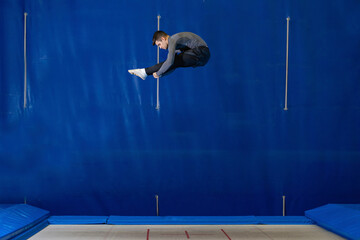 Young man doing an acrobatic figure on elastic bed, in blue background