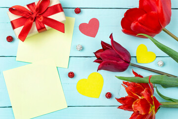 The concept of the celebration. Red tulips, a gift in a box, beads, hearts and yellow sheets of writing paper on a blue (mint) background. Space for text. Valentine's day, women's day, birthday.