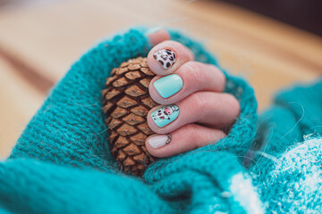 Hand with fresh manicure holding a pine cone. Playful nail design for summer, hand wrapped in a handmade knitted sweater. Selective focus on the details, blurred background.