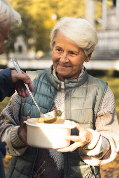 Wholesome caucasian grandmother shares her homemade soup with homeless people in park. Helping people in need. Vertical Shot. High quality photo