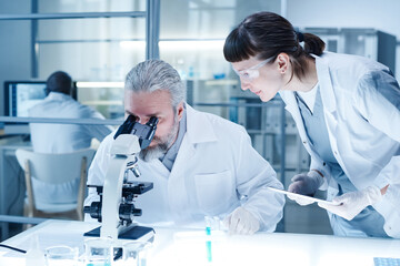 Mature bearded man in white coat examining analysis through the microscope with woman entering data...