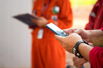 A worker is using smartphone to searching some information during safety meeting with other colleague (as blurred background). Industrial worker action photo. Close-up and selective focus at hand.