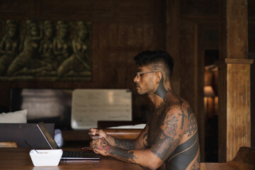Fototapeta na wymiar Hispanic tattoed young man working at the laptop in outdoor kitchen at private villa while having a breakfast