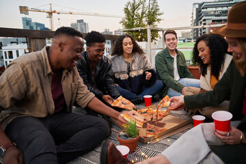 Group of diverse friends grabbing pieces of pizza to eat while sitting on rooftop terrace in the city