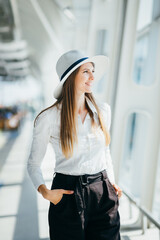 Young woman wearing casual clothes is posing at airport and waiting near luggage in hall airplane departure