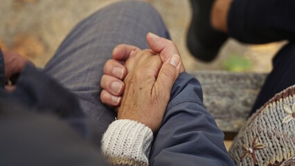 Caucasian elderly couple holding their old, wrinkled hands together while sitting on a bench. High quality photo