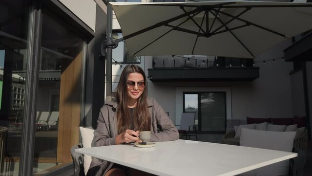 Gorgeous young woman in trendy sunglasses and outfit sitting on outdoors terrace and drinking freshly brewed coffee. Female tourist enjoying relaxing time at modern hotel.