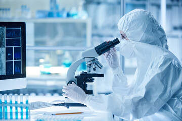 Female scientist in protective wear examining the sample of new virus through the microscope sitting at table in laboratory - obrazy, fototapety, plakaty