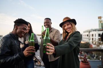 Group of multi-cultural young adults making a toast with drinks on a rooftop terrace in the city. 