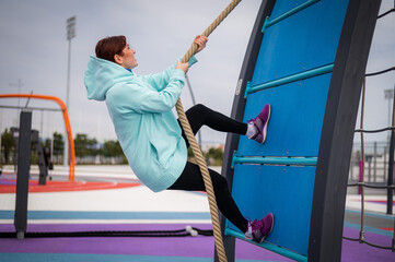 Caucasian woman in a mint sweatshirt climbing a tightrope at an outdoor sports ground. 