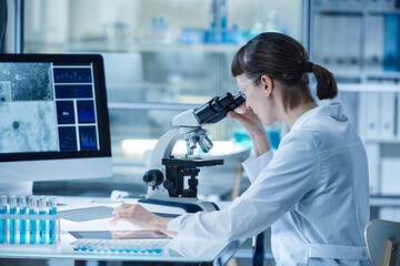 Young woman in lab coat looking through the microscope while sitting at table with computer and test tubes during her work in laboratory - obrazy, fototapety, plakaty