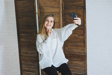 Emotional young woman in a white cotton shirt gesticulates and looks at the screen of a modern...